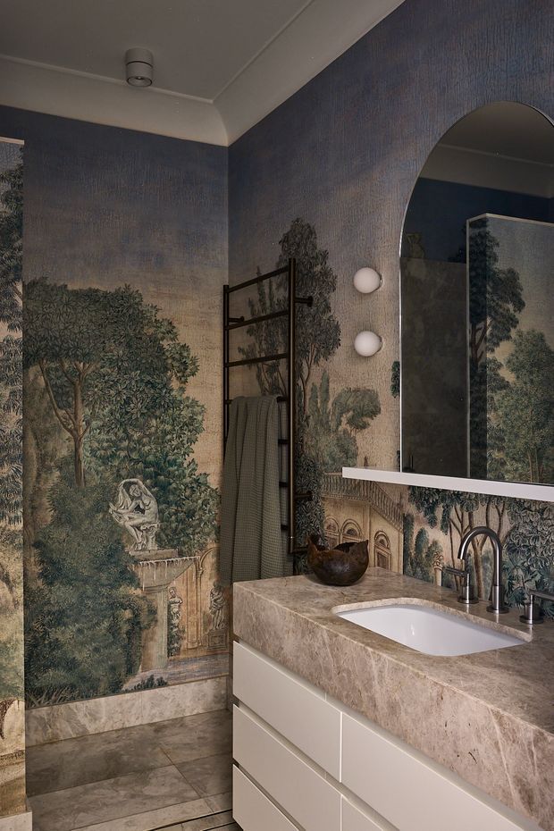 A moody, richly decorated bathroom featuring a large classical woodland mural wallpaper, an arched wall opening with mirror, twin globe wall lights, a black ladder-style towel rail, and a pale stone vanity with integrated basin and brushed metal tapware. Large-format stone-look floor tiles and warm neutral cabinetry complete the refined interior.