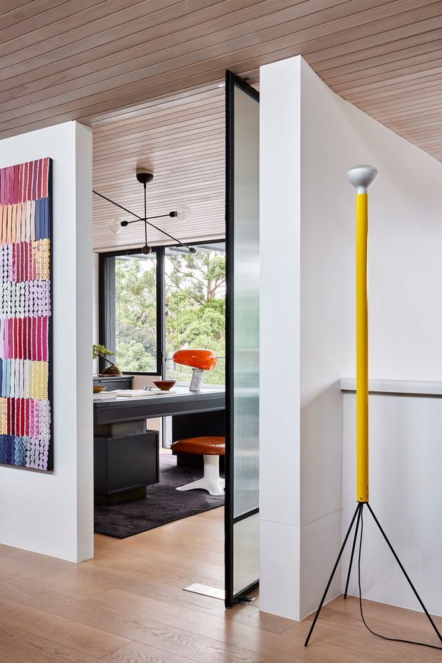 Bright contemporary interior view looking through a partially open black-framed glazed partition into a home office. The space features white walls, warm light timber flooring and a slatted timber ceiling, with a colourful textile wall hanging in the foreground. The office contains a dark built-in desk, orange sculptural chair, large window, and modern globe-style chandelier. A tall yellow tripod floor lamp adds a strong accent in the foreground.