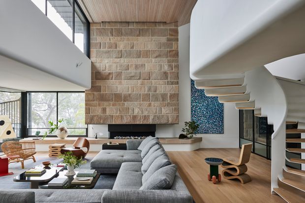 A spacious, double-height contemporary living room with a large grey sectional sofa arranged around a low coffee table. The room features a textured natural-stone fireplace wall, a linear fireplace, warm timber ceiling and flooring, expansive black-framed glazing, a sculptural white stair, and a colourful abstract wall artwork. Curved architectural forms and carefully selected mid-century-inspired furniture create a refined, modern interior.