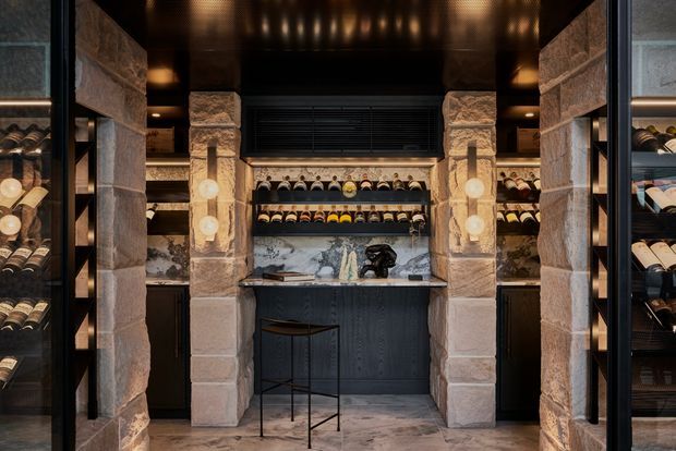 A dramatic, compact wine room or home bar framed by chunky natural stone piers. Dark built-in wine racks line both sides, while a central black bar cabinet and stool sit beneath a wine display and wall-mounted cooling unit. The ceiling and cabinetry are black, with warm recessed lighting and a marble-like stone backsplash and floor creating a luxurious, moody atmosphere.