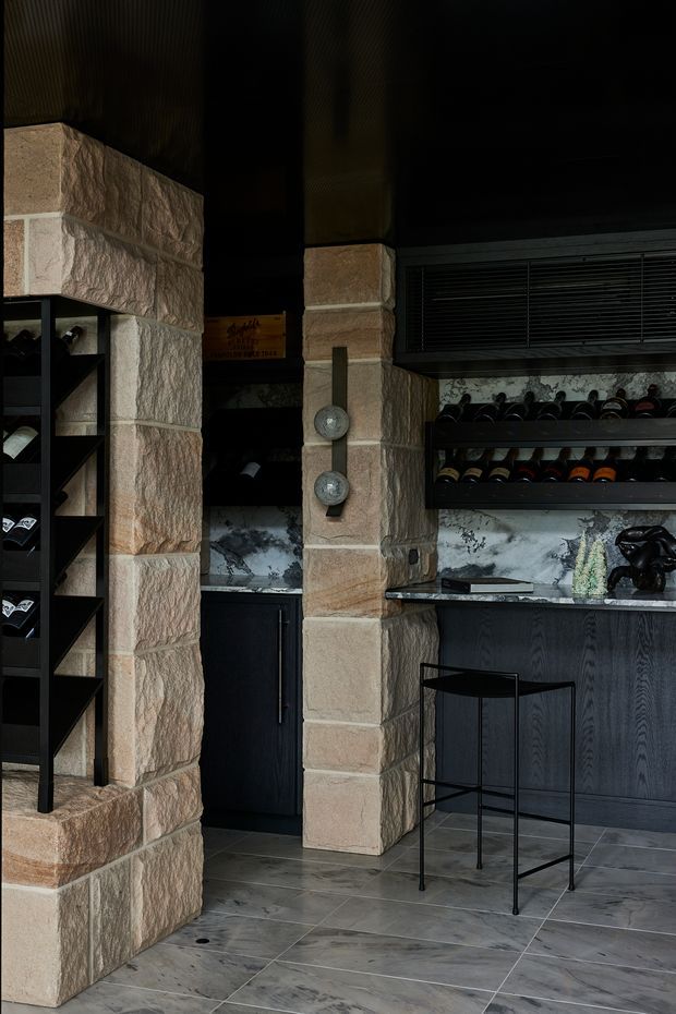 A dark, refined wine storage and bar area featuring substantial textured beige stone piers, black cabinetry, open wine racks, a marble-look backsplash and benchtop, dark ceiling, and large grey stone-look floor tiles. A slim black bar stool and decorative objects complete the moody interior.