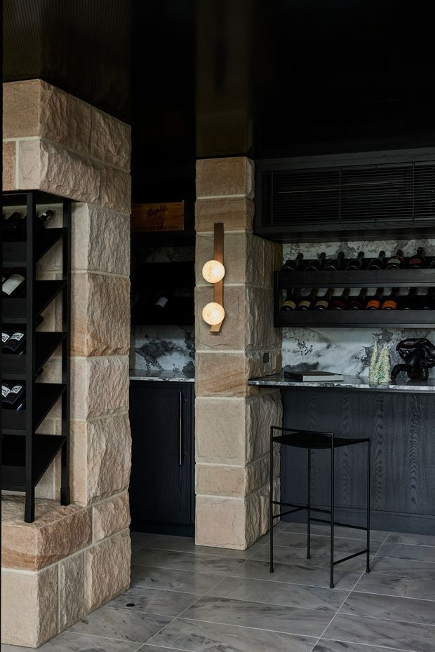 A dark, refined wine storage and serving area with rough-cut beige stone columns and shelving, black cabinetry, grey stone-look tiled flooring, a marble-patterned backsplash, wall-mounted globe lights, and extensive bottle racks. The space has a dramatic, intimate atmosphere with strong natural-textured masonry and black joinery.