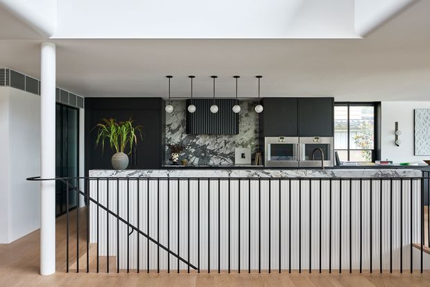Contemporary open-plan kitchen viewed from the living area, featuring a large island with vertically fluted light cabinetry and a slim black metal balustrade, dark upper cabinetry, a dramatic veined stone splashback, integrated appliances, pendant lighting, and warm timber flooring. The kitchen opens onto a bright, high-ceilinged space with large windows and a sculptural white column.