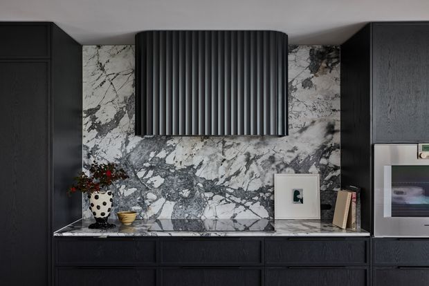 A dramatic, dark-toned contemporary kitchen wall featuring black textured cabinetry, a striking veined marble splashback and benchtop, and a sculptural ribbed black rangehood. Integrated appliances and minimal decorative objects complete the refined, high-contrast composition.