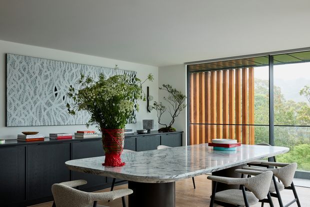 Contemporary dining area with a large polished stone dining table surrounded by upholstered chairs. A dark built-in sideboard runs along the wall beneath a patterned feature artwork, while floor-to-ceiling glazing and vertical timber screening open to a leafy outlook. The room is styled with sculptural greenery and restrained modern furnishings.
