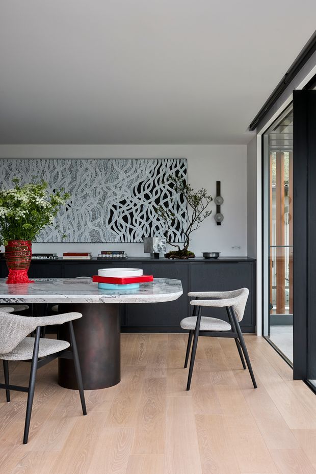 Contemporary dining and living space with a round marble dining table, sculptural dark cylindrical pedestal, upholstered black-framed dining chairs, dark built-in cabinetry, light timber flooring, and a large monochrome abstract artwork. Full-height glazed doors open to an exterior balcony or terrace, bringing in natural light.