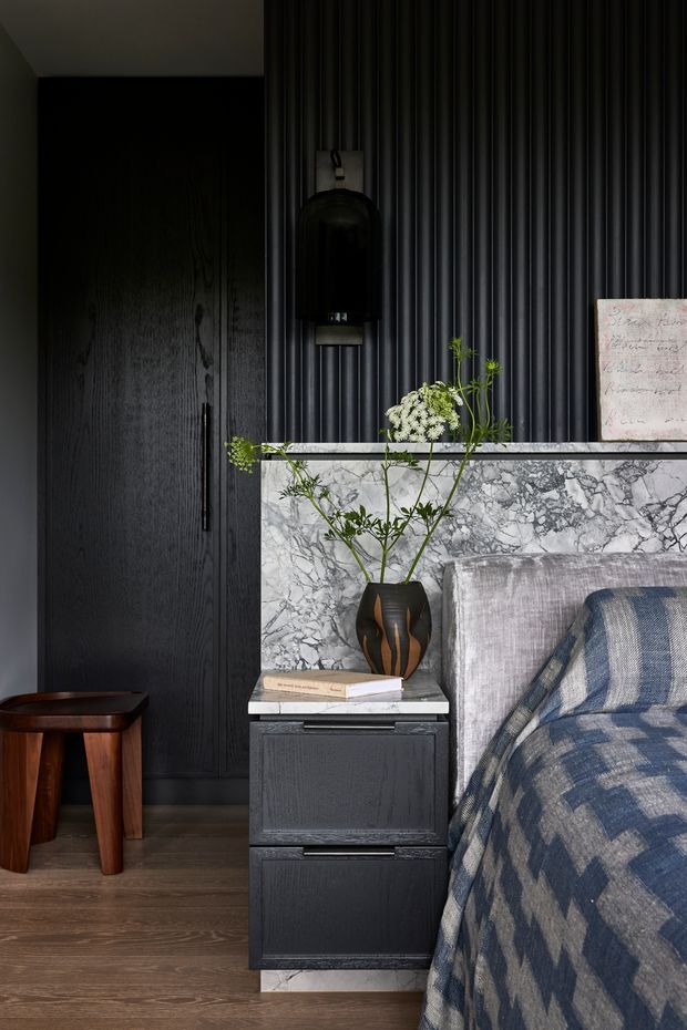 A moody, refined bedroom vignette featuring dark vertical fluted wall panelling, a black timber door and built-in bedside cabinetry. A grey-and-white veined stone headboard wall and bedside surface contrast with the dark joinery. The bed is dressed in a blue, grey and cream geometric textile, with a sculptural dark vase holding delicate greenery, books, and a black wall-mounted pendant light. Dark timber flooring and a small wooden stool complete the composition.