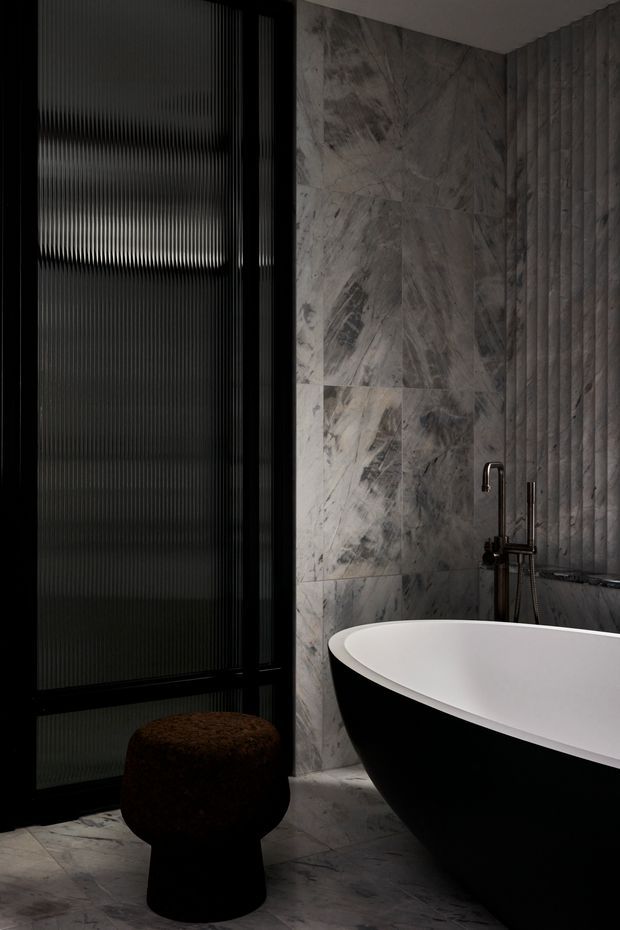 A dark, luxurious bathroom featuring a freestanding black-and-white bathtub, a black freestanding bath tap, large veined grey marble wall and floor tiles, and a vertically ribbed textured wall. A black-framed fluted-glass window or shower screen occupies the left side, with a small dark upholstered stool in the foreground. The space has a moody, refined, high-contrast atmosphere.