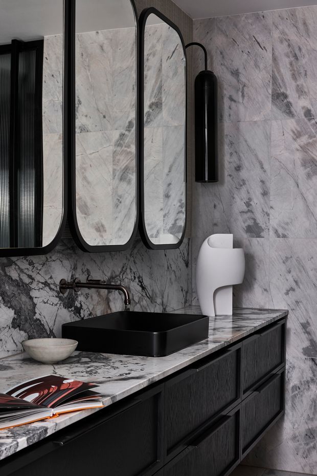 A refined, dark-toned bathroom vanity featuring a large veined marble-look stone wall and benchtop, matte black rectangular vessel basin, black wall-mounted tapware, sculptural black-framed mirrors, black cabinetry, and a minimalist black wall light. A white sculptural object and small bowl sit on the counter, adding contrast.