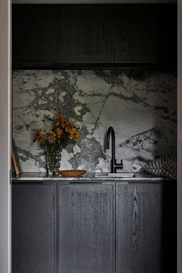 A dark, moody compact kitchen or bar-style kitchenette with charcoal-black oak cabinetry, a dramatic veined grey-and-white natural stone backsplash and benchtop, and a black gooseneck mixer tap. A vase of flowers, wooden bowl, books and patterned cushion add understated styling.