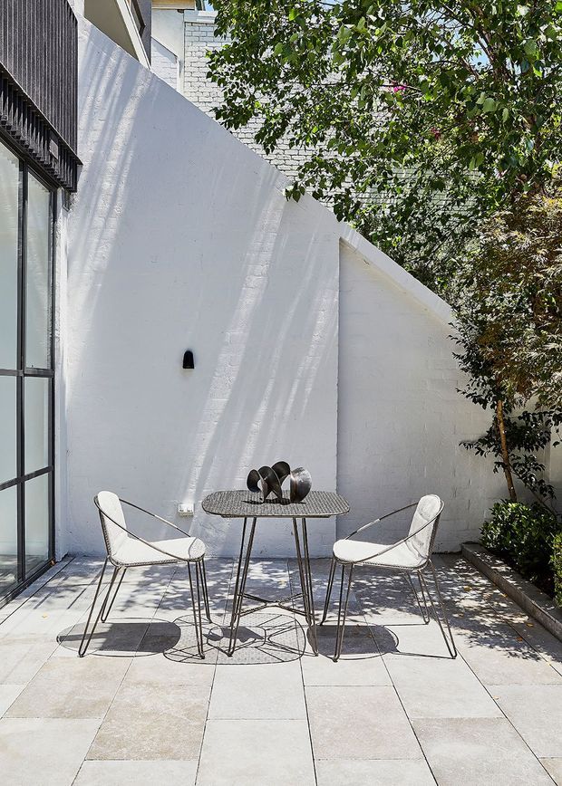 A bright, compact courtyard or patio with white painted masonry walls, large pale paving slabs, and a small round outdoor dining table with two wire-frame chairs. A glazed door or window with dark metal framing opens onto the space, while established trees and planting provide dappled shade. The setting has a clean, minimalist contemporary character.