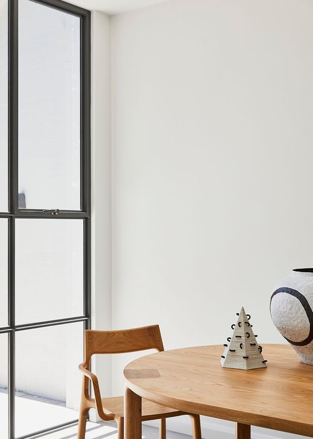 A bright, minimalist dining area with a round light timber dining table and matching sculptural timber chair beside a large black-framed window. A simple white wall forms the backdrop, with understated decorative objects on the table. The space has clean lines, abundant natural light, and a restrained contemporary aesthetic.