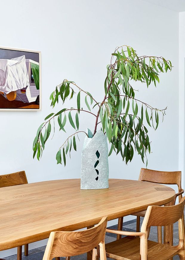 Bright dining room featuring a large oval solid timber dining table with matching sculptural timber chairs. A tall textured ceramic vase with leafy branches forms the centrepiece, while a framed artwork hangs on the pale wall. The room has a clean, contemporary and natural aesthetic.