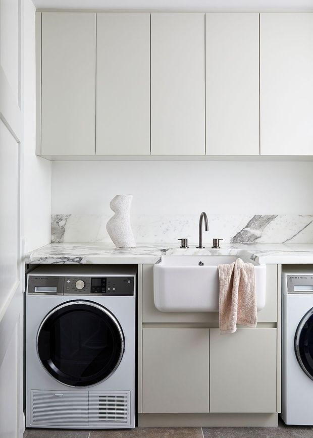 Bright, compact contemporary laundry with white built-in cabinetry, a marble-look benchtop and splashback, farmhouse-style undermount sink, chrome tapware, and front-loading washer and dryer integrated beneath the counter. The room has a clean, minimalist aesthetic with pale neutral finishes and soft blush towels.