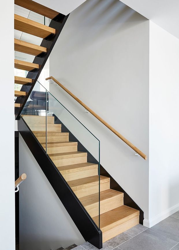 A bright contemporary stairway with open-riser timber treads, black steel stringers, a clear glass balustrade, and a timber handrail. White walls and grey floor tiles create a clean, minimalist interior, with an upper-level landing and additional open stair flight visible above.