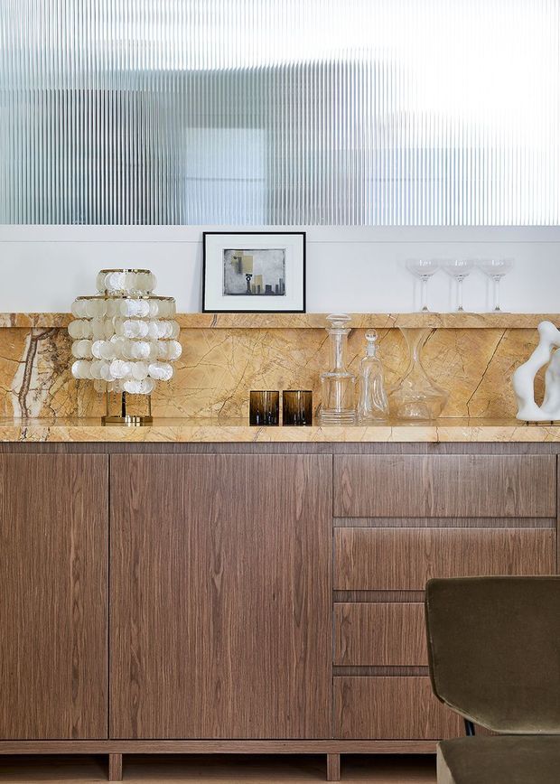 Close-up interior view of a refined built-in timber sideboard or cabinet, featuring vertically grained dark wood cabinetry on short legs, a striking veined amber stone shelf and backsplash, and a ribbed translucent upper wall panel. Decorative glassware, a sculptural lamp, framed artwork, and a partial upholstered chair add a luxe, curated character.