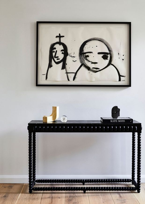 A styled interior vignette featuring a narrow black console table with ornate bead-like detailing and turned legs against a pale wall. The console displays sculptural objects and books, with a large black-framed monochrome artwork hung above. Light timber flooring and restrained black, white, and natural wood tones create a refined, contemporary setting.