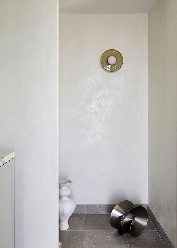 A minimalist interior alcove with softly textured pale plaster walls, dark grey tiled flooring and skirting, and a small circular metallic wall light or feature. Sculptural white and dark metallic decorative objects sit on the floor, creating a restrained gallery-like composition.