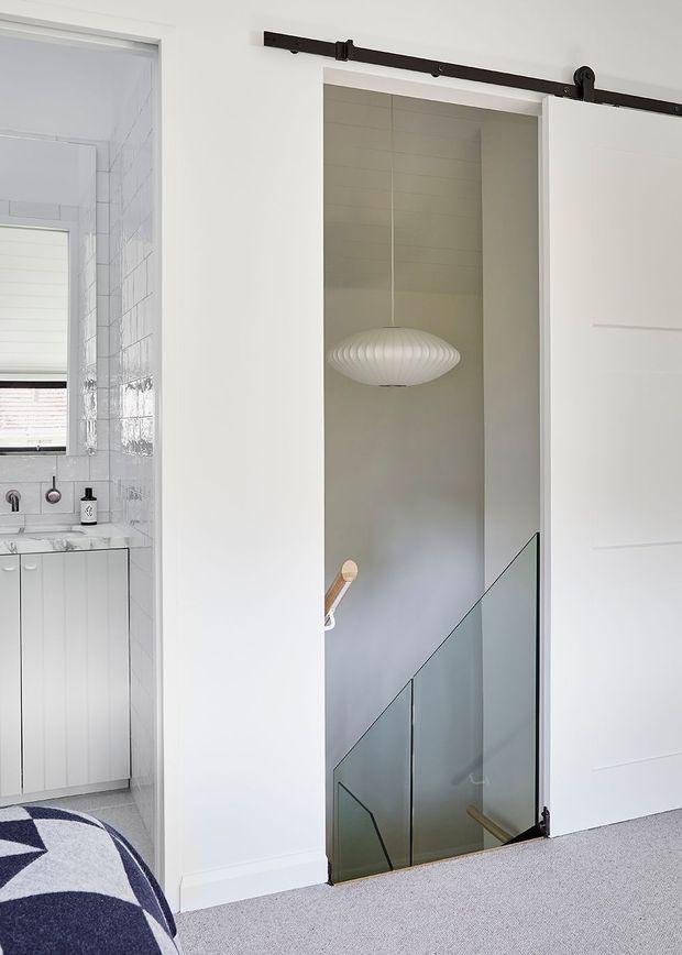 Bright contemporary residential interior showing a hallway and stairwell behind a white barn-style sliding door. The stairwell has pale grey walls, a suspended sculptural pendant light, timber handrail, and frameless glass balustrade. A bathroom with white tiled walls, marble-look vanity, and white cabinetry is visible to the left, while part of a bed and patterned rug appear in the foreground.