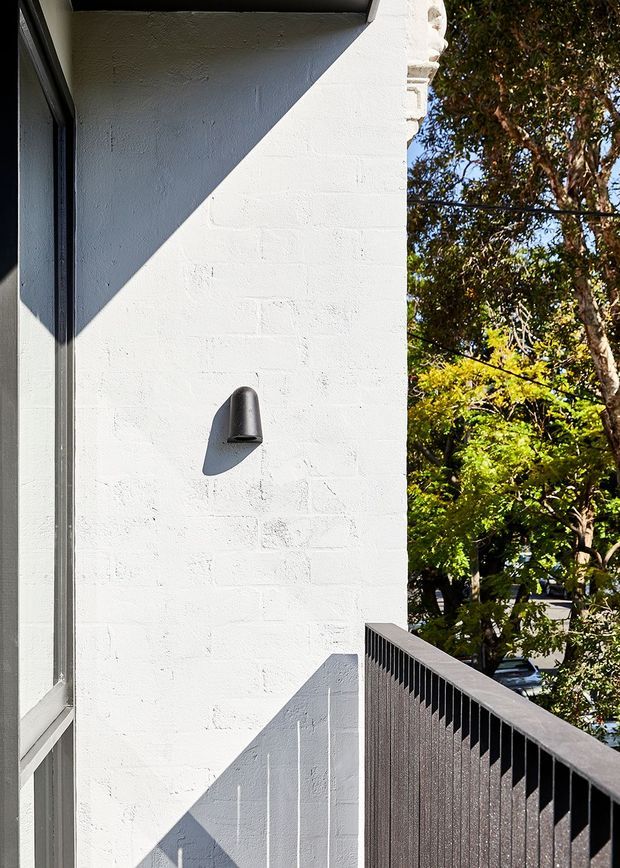 Close-up exterior view of a white-painted textured masonry wall with a dark wall light, narrow window frame, and dark vertical-slat balcony or terrace balustrade. Strong sunlight creates angular shadows across the facade, with leafy trees and a suburban street visible beyond.