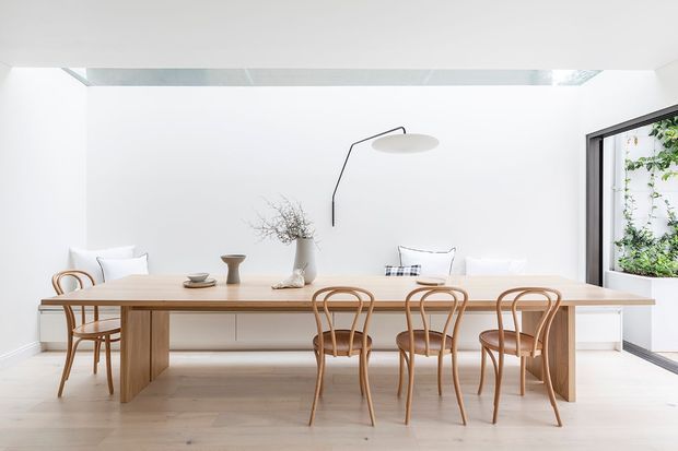 Bright minimalist dining area with a long light timber dining table and matching bentwood chairs. White walls, pale timber flooring, a slim black wall-mounted pendant, low built-in cabinetry, and a large glazed opening with greenery create an airy, restrained interior.