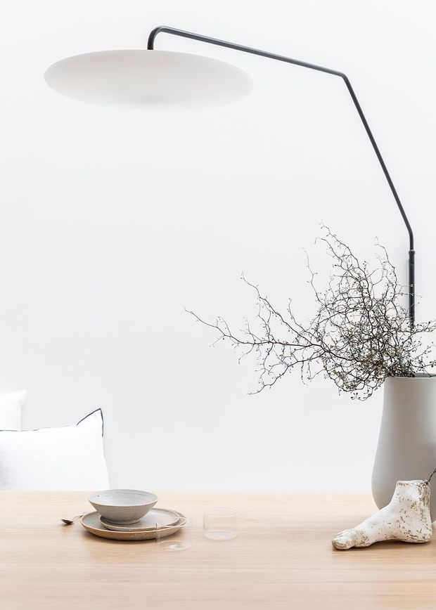 Minimalist product-style interior vignette featuring a large arc floor lamp with a slim dark frame and broad white shade, positioned behind a pale timber dining table. The table is styled with neutral ceramic tableware, clear glassware, a tall matte vase containing bare branches, and a sculptural light-toned object. The background is plain white, creating a calm, refined Scandinavian-inspired composition.
