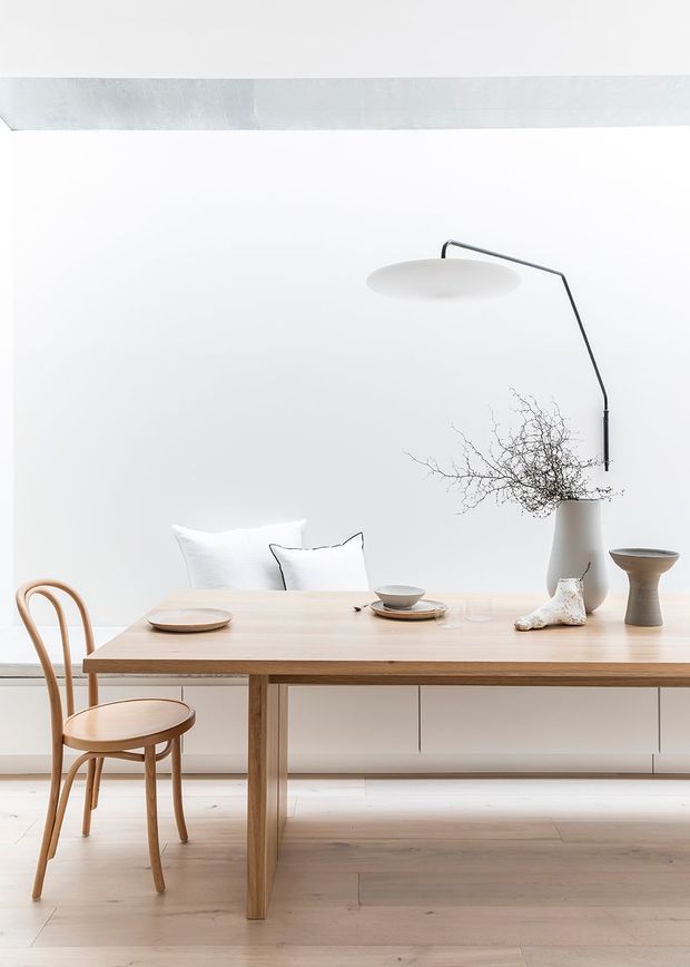 A bright, minimalist dining area with a long light timber dining table, a curved bentwood chair, white upholstered seating, and a large black arched wall lamp. A simple ceramic vase with bare branches, tableware, and sculptural objects add restrained decorative detail. The room has white walls, pale timber flooring, and low built-in cabinetry beneath the wall.
