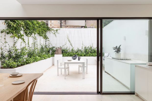 Bright contemporary interior opening directly onto a small landscaped courtyard. The image shows a pale timber dining table and chairs in the foreground, wide dark-framed glazed sliding doors, a white outdoor table and chairs, planted garden beds, and a minimal white kitchen or joinery area visible on the right.