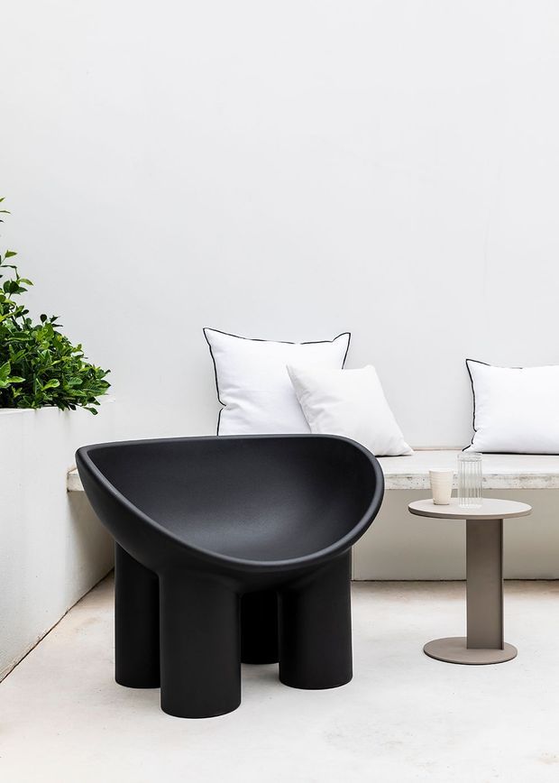 A minimalist seating vignette featuring a sculptural matte-black lounge chair with a rounded, enveloping form, positioned beside a small round side table. White cushions rest on a built-in white bench against a plain white wall, with a leafy potted plant adding greenery at the left. The palette is monochromatic and the composition is clean and contemporary.