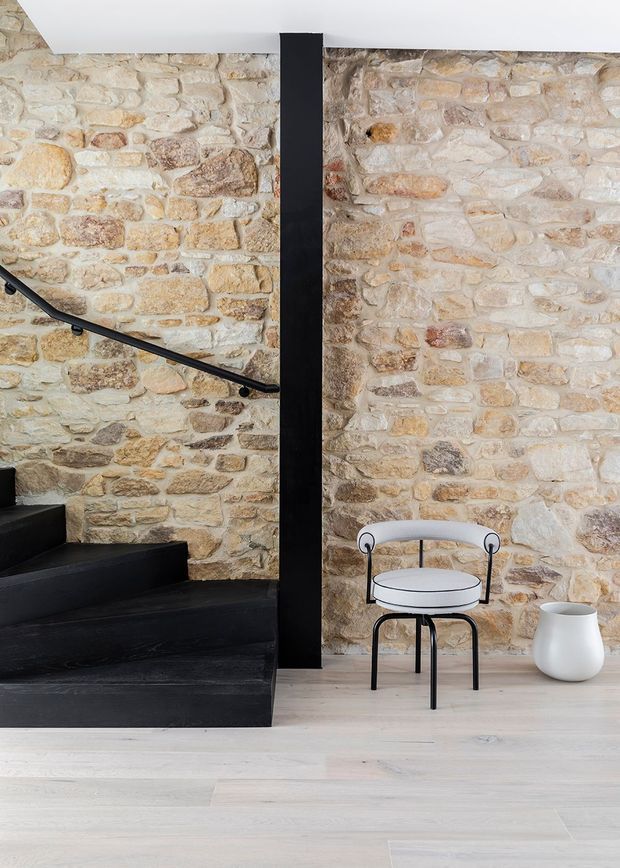A refined interior stairwell featuring a full-height exposed natural stone wall, black-painted staircase and central structural post, and a minimalist black metal handrail. A small round side table and white ceramic vessel sit on pale timber flooring, creating a strong contrast between rustic masonry and contemporary detailing.