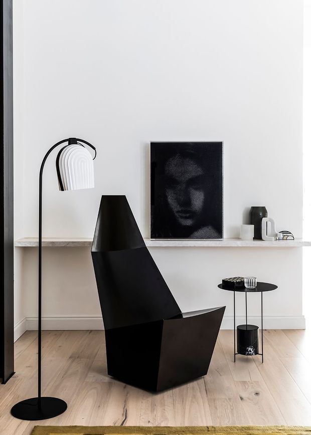 A refined minimalist living room with white walls, pale timber flooring, and a narrow stone or concrete shelf running along the wall. The room is furnished with a sculptural black angular lounge chair, a slender black arched floor lamp with a white ribbed shade, a small round black side table, and monochrome artwork and decorative objects. The restrained black-and-white palette and sculptural furniture create a sophisticated contemporary setting.