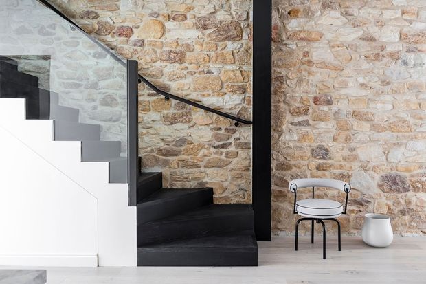 A minimalist stairway with dark timber or painted treads and black metal handrails, set against exposed irregular stone masonry walls. The space includes a simple black-framed chair and white ceramic vessel, with pale flooring and a bright, restrained palette.