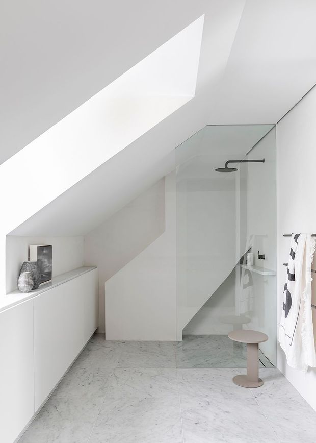 A bright, minimalist bathroom beneath a sloping ceiling, featuring a frameless glass walk-in shower with a matte black shower fixture, pale marble-look floor tiles, white built-in cabinetry, and a small round stool. A skylight-like roof opening brings in strong natural light.