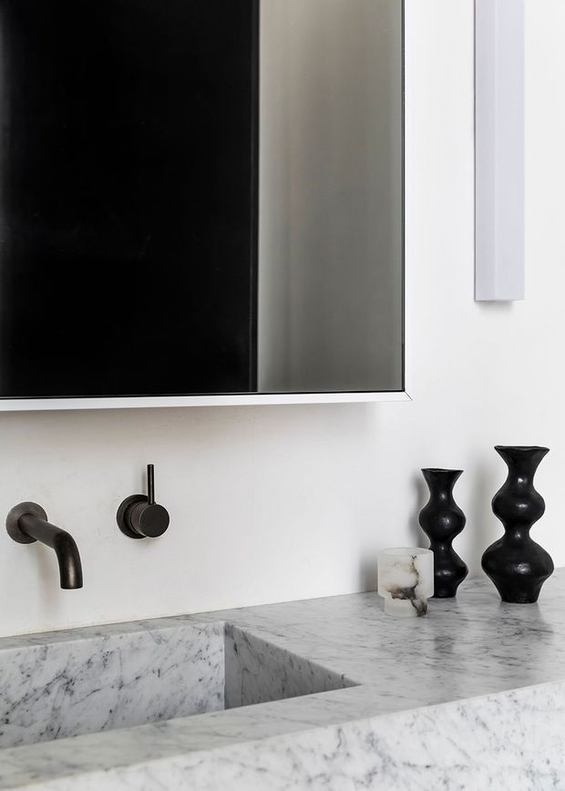 Close-up of a refined minimalist bathroom vanity featuring a pale veined marble benchtop and integrated trough basin, dark wall-mounted tapware, a large frameless mirror, and sculptural black decorative vessels.