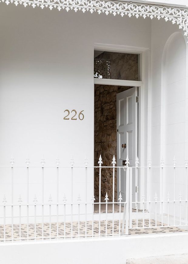 A close-up exterior view of a white-painted heritage-style porch and entryway. Decorative wrought-iron fretwork runs along the roof edge and balustrade, with a white panelled front door opening into a stone-lined entry. The house number 226 is mounted beside the door, and patterned tile paving is visible underfoot.