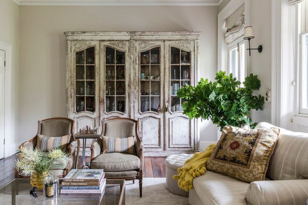 Elegant traditional living room with a distressed antique-style glass-front cabinet, two upholstered armchairs, a neutral sofa, a glass coffee table, and layered decorative textiles. The room has pale beige walls, white trim, tall windows with Roman shades, and dark timber flooring, creating a refined, warm, classic interior.