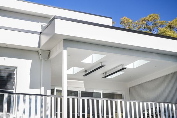 Bright exterior view of a contemporary white flat-roofed house, focused on a covered outdoor terrace or balcony with slim metal balustrades, skylights, linear ceiling-mounted lighting, and a ribbed vertical feature wall. Clean geometric volumes and dark roof trims contrast with the white facade, with trees visible beyond.
