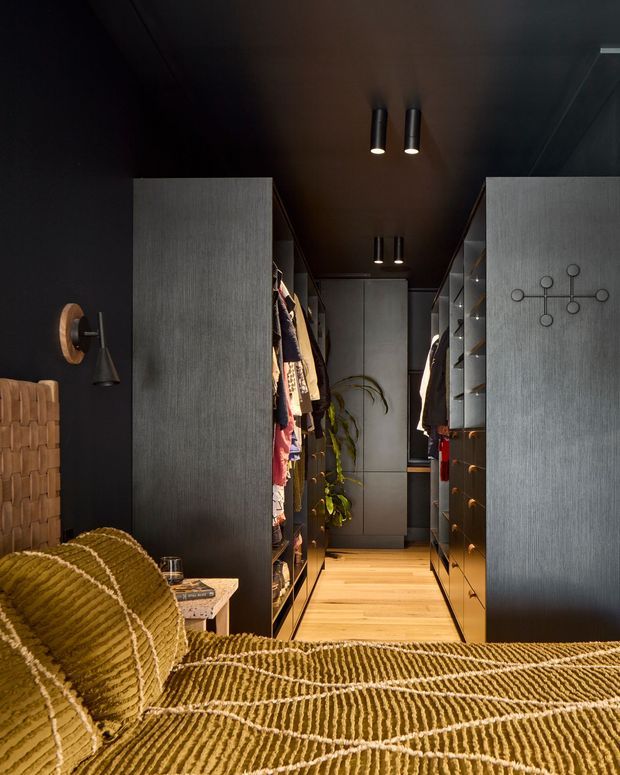 A moody bedroom with a walk-in wardrobe visible directly beyond the bed. The room features dark charcoal walls and ceiling, a textured woven headboard, mustard-gold patterned bedding, warm timber flooring, and extensive dark grey built-in wardrobe cabinetry with open shelving and hanging clothes. Slim cylindrical pendant and wall lights provide focused illumination, while a plant adds a small natural element at the rear of the wardrobe.