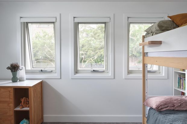 Bright compact bedroom with three tall white-framed windows, a timber storage cabinet with open shelving, and a timber bunk or loft bed with bedding and integrated shelving. The room has pale grey walls, white trim, and a simple, functional contemporary fit-out.