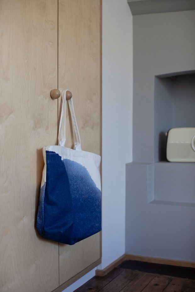 Close-up interior view of a compact entryway featuring tall, light timber built-in cabinetry with round wooden hooks and a hanging blue-and-white tote bag. A recessed wall niche with a small retro radio is visible to the right. The space has muted grey walls, dark timber flooring, and simple understated detailing.