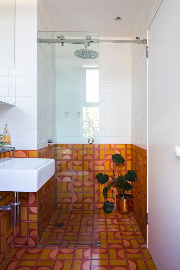 A compact, brightly styled bathroom with a walk-in shower enclosed by clear glass and a brushed metal sliding door rail. The walls and floor are clad in vivid retro geometric tiles in orange, terracotta, mustard yellow, brown, and purple tones, with white rectangular wall tiles above. A white wall-mounted basin is visible at left, and a potted plant adds greenery near the shower.