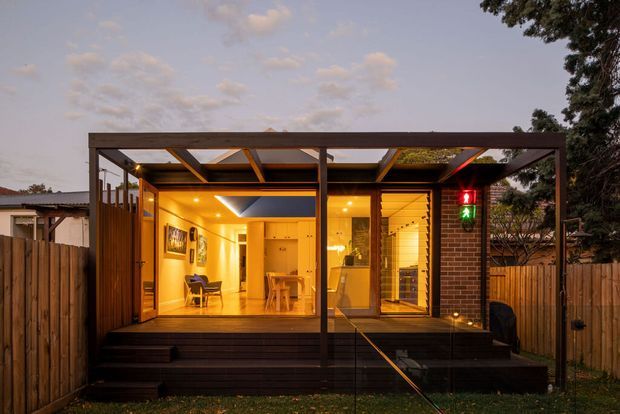 A compact modern timber-framed extension or outdoor pavilion viewed from the garden at dusk. The open glazed facade reveals a warmly lit interior with a small dining area, kitchenette or living space, timber floors and walls, and a distinctive angular ceiling skylight. Dark timber decking, steps, vertical timber fencing, exposed framing and a partially transparent roof create a sheltered indoor-outdoor connection.