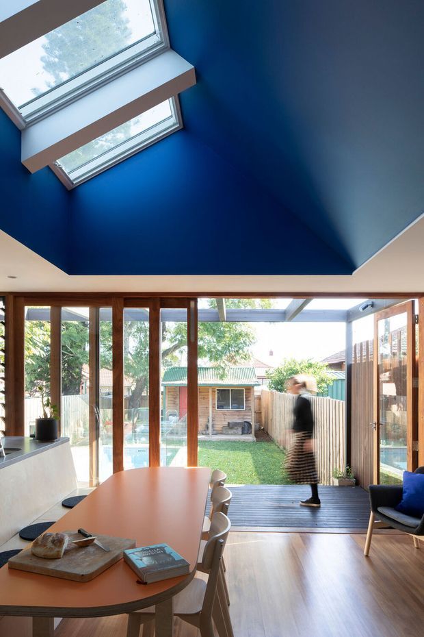 Bright open-plan dining and living area with a large timber-framed glazed opening to a private backyard. The room features a warm timber floor, a rounded orange dining table with chairs, a compact kitchen bench at left, and a dramatic deep-blue angled ceiling with skylights. A timber deck and garden are visible beyond the glazing.