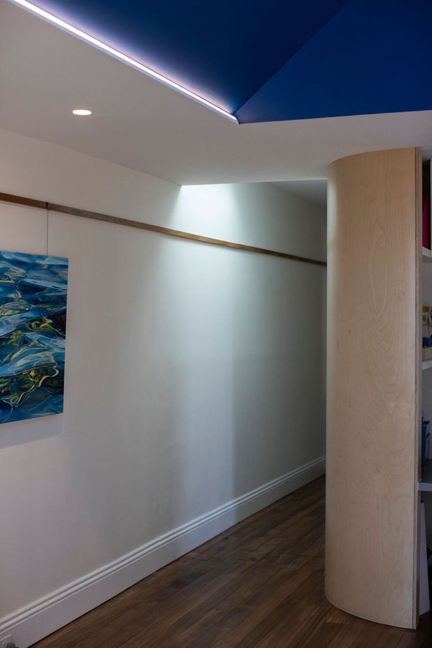 A compact interior hallway with white walls, dark timber flooring, white skirting boards, and a curved light-wood built-in partition or shelving unit. A vivid abstract artwork hangs on the wall, while a dramatic dark-blue recessed or angled ceiling feature with concealed LED lighting creates a strong architectural focal point.