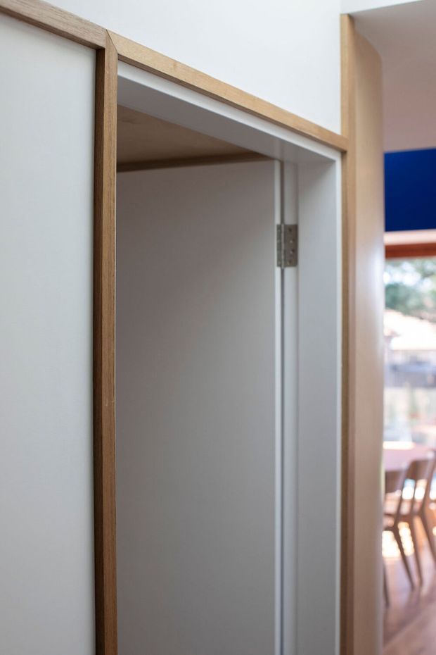 Close-up architectural detail of a white interior door or built-in opening with exposed light timber edging, visible metal hinge hardware, and a glimpse of a bright dining or living area beyond. The image emphasizes precise joinery and contrasting blue wall or artwork in the background.