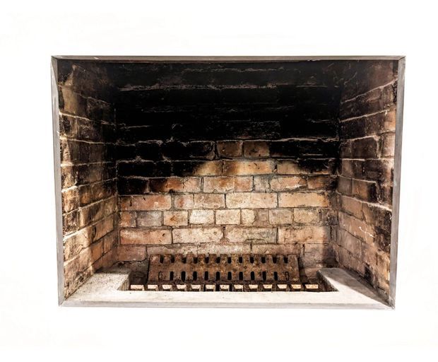 Close-up view of an old masonry fireplace or hearth, with heavily soot-darkened brickwork, a metal grate, and a pale stone or rendered surround against a plain white background.