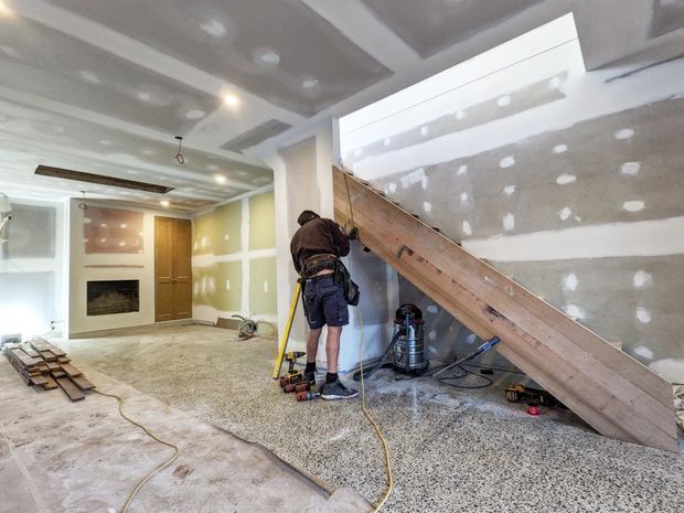 Interior construction scene showing a partially renovated open-plan room with a timber staircase, unfinished drywall ceilings and walls, exposed electrical work, tools, and a worker. A fireplace, door, and rough flooring are visible in the background.