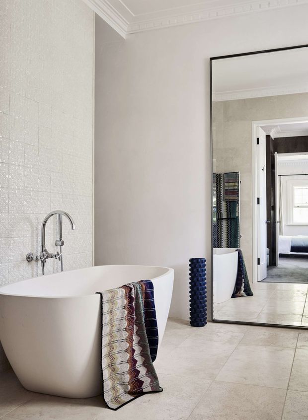 Bright, refined bathroom featuring a freestanding white oval bathtub, wall-mounted chrome tapware, textured white tiled feature wall, pale stone-look floor tiles, decorative ceiling cornice, and a large black-framed leaning mirror. The mirror reflects an adjoining bedroom and bathroom furnishings. A patterned throw hangs over the tub, with a tall dark decorative object beside the mirror.