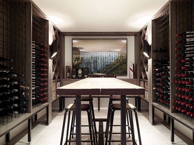 A refined wine room with extensive floor-to-ceiling wine bottle racks lining both side walls. A large central island with a pale stone-like top and dark timber base provides standing and seated tasting space, with stools tucked beneath. Built-in timber shelving, integrated lighting, and a framed vineyard view or artwork complete the space.
