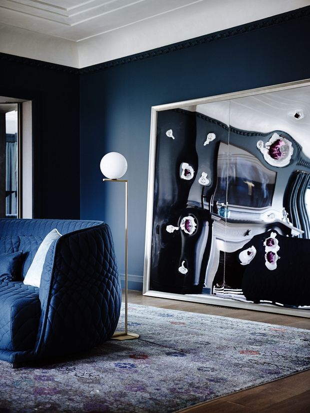 A sophisticated living room with deep navy walls, ornate white ceiling detailing and dark decorative cornice trim. A plush quilted navy sofa occupies the foreground, accompanied by a slim brass-and-white globe floor lamp. A large framed abstract artwork leans against the wall, while a muted patterned area rug covers much of the timber floor. The room has a dramatic, luxurious and refined atmosphere with a strong dark-blue and white palette.